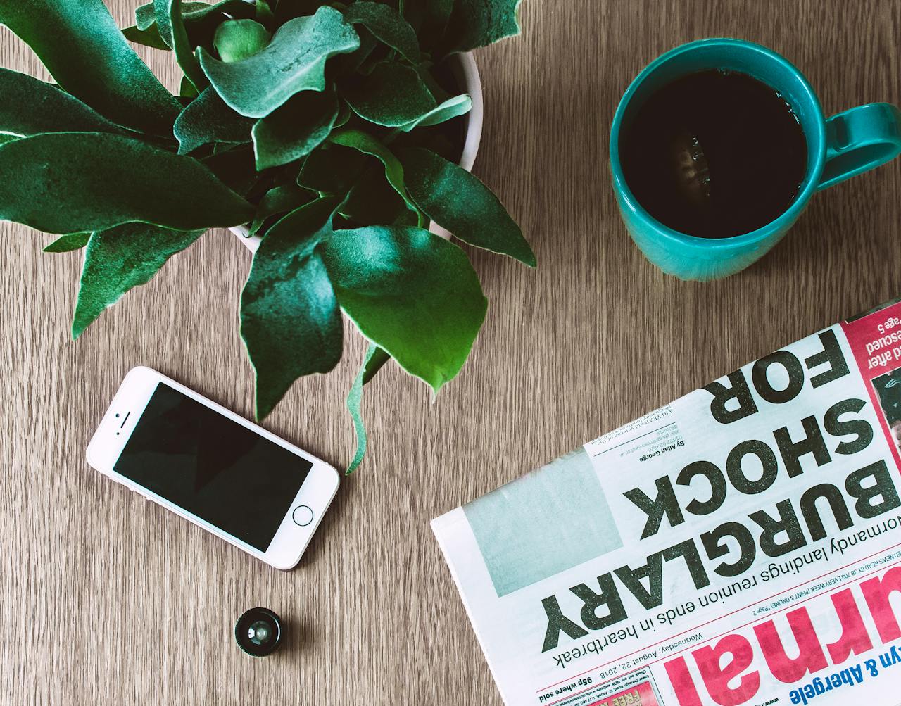 Flat lay arrangement of a coffee mug, newspaper, smartphone, and plant on a wooden table.